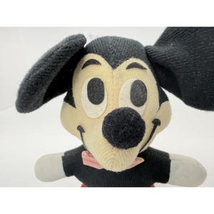 Puede incluir: Un peluche vintage de Mickey Mouse. El peluche presenta un cuerpo, orejas y nariz negros, con una cara color crema y una pajarita rosa. El juguete tiene ojos negros grandes y una sonrisa cosida. El peluche est&aacute; hecho de un material suave y texturizado.