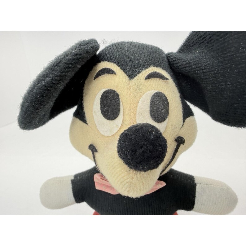 Puede incluir: Un peluche vintage de Mickey Mouse. El peluche presenta un cuerpo, orejas y nariz negros, con una cara color crema y una pajarita rosa. El juguete tiene ojos negros grandes y una sonrisa cosida. El peluche est&aacute; hecho de un material suave y texturizado.