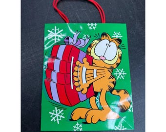 May include: A green gift bag featuring a cartoon illustration of Garfield the cat holding a large red and purple gift box with a purple bow. The bag is decorated with white snowflake patterns and has two red cord handles at the top for carrying.