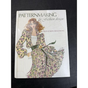 May include: A hardcover book titled Patternmaking for Fashion Design by Helen Joseph Armstrong. The white cover features a detailed fashion illustration of a person wearing a long-sleeved, floral-patterned dress with a green waist sash.