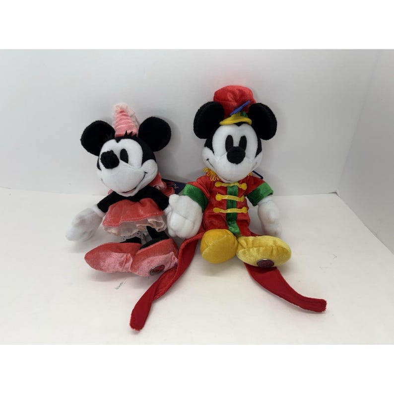 Puede incluir: Dos mu&ntilde;ecos de peluche de personajes de Disney. Uno es Minnie Mouse, con un atuendo rosa y rojo con un lazo a juego. El otro es Mickey Mouse, vestido con un atuendo rojo y dorado de estilo circo con un sombrero rojo. Ambos tienen orejas negras.