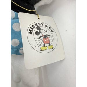 K&ouml;nnte beinhalten: Ein wei&szlig;es Etikett mit dem Logo "MICKEY & CO." mit Mickey Mouse in einem Kreis. Das Etikett ist an einem Pl&uuml;schtier mit einem blauen Polka-Dot-Kleid befestigt.