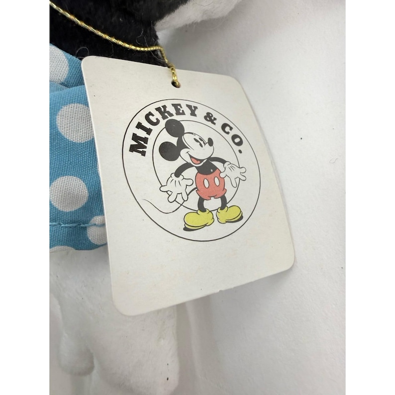K&ouml;nnte beinhalten: Ein wei&szlig;es Etikett mit dem Logo "MICKEY & CO." mit Mickey Mouse in einem Kreis. Das Etikett ist an einem Pl&uuml;schtier mit einem blauen Polka-Dot-Kleid befestigt.