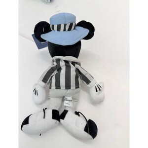 May include: A plush Mickey Mouse toy wearing a light blue hat with black and white stripes, a black and white striped shirt, and white gloves. The toy has black ears and black and white shoes. The Disney logo is visible on the toy.