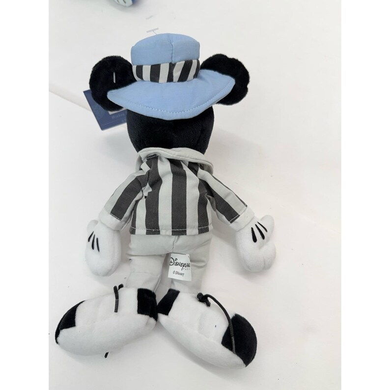 May include: A plush Mickey Mouse toy wearing a light blue hat with black and white stripes, a black and white striped shirt, and white gloves. The toy has black ears and black and white shoes. The Disney logo is visible on the toy.