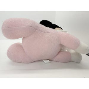 Puede incluir: Un peluche en posici&oacute;n tumbada, con cuerpo rosa claro, cabeza negra y detalles blancos. El juguete tiene una textura suave y difusa y est&aacute; dise&ntilde;ado para parecerse a un personaje. El juguete est&aacute; sobre una superficie blanca.