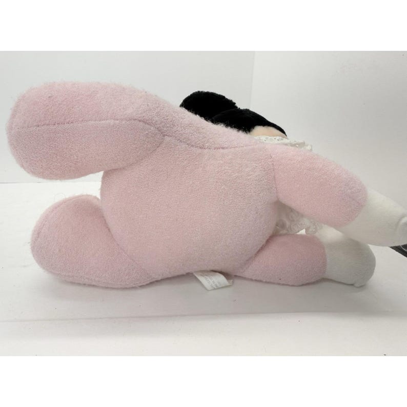 Puede incluir: Un peluche en posici&oacute;n tumbada, con cuerpo rosa claro, cabeza negra y detalles blancos. El juguete tiene una textura suave y difusa y est&aacute; dise&ntilde;ado para parecerse a un personaje. El juguete est&aacute; sobre una superficie blanca.