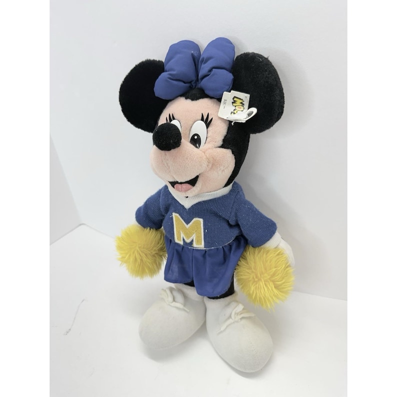 May include: A Minnie Mouse plush doll wearing a blue outfit with a gold "M" on the chest, yellow pom-poms, and white shoes. The doll has a blue bow and black ears. A tag is attached to the doll.
