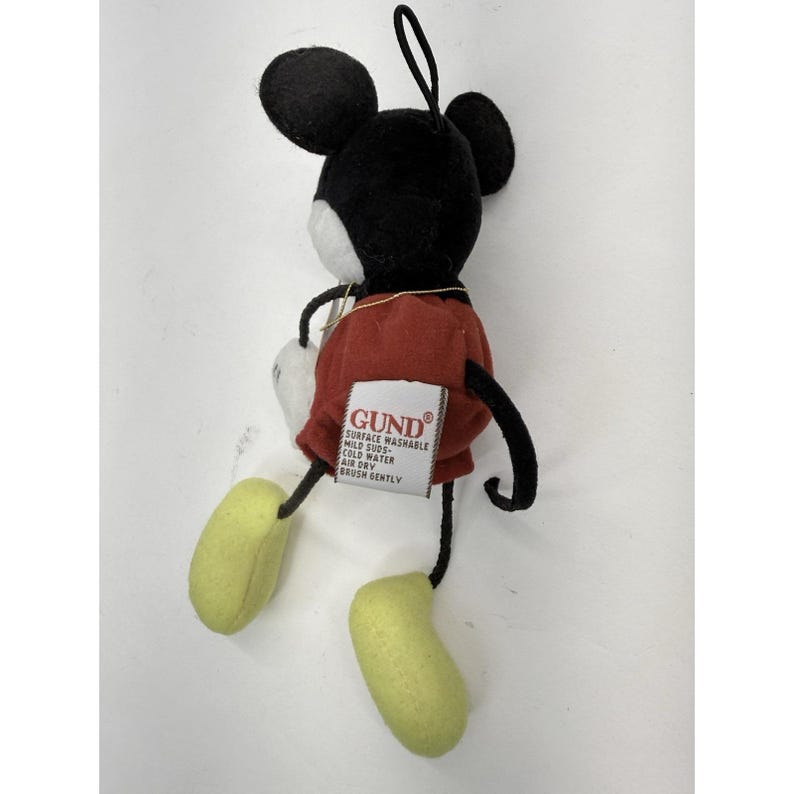 K&ouml;nnte beinhalten: Ein Pl&uuml;schtier von Mickey Mouse mit schwarzen Ohren, einem schwarzen Kopf, einem wei&szlig;en Gesicht, roten Shorts und gelben F&uuml;&szlig;en. Das Spielzeug hat eine schwarze Schlaufe zum Aufh&auml;ngen und ein wei&szlig;es Etikett mit der Marke "GUND".