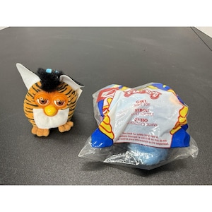 May include: A plush toy with orange and black tiger stripes, a white belly, and black hair tufts, alongside a second plush toy sealed in a clear plastic bag with blue, yellow, and red packaging featuring the text Owl Soft Toy and Hibou Jouet Souple.