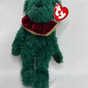 May include: Green plush beanie baby bear with a red velvet collar and gold trim. The bear has a black nose and black eyes. The bear has a Ty tag attached to its ear.