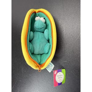 May include: A plush teal frog toy nestled inside a yellow, oval-shaped zippered pouch with a green interior. An orange zipper runs along the edge. A tag attached to the pouch features the text Sunshine Softie and a small suitcase icon.