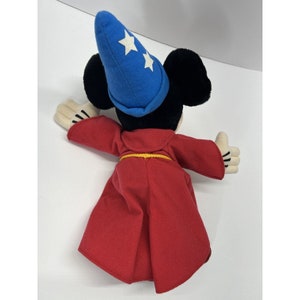 May include: A plush toy of Mickey Mouse dressed as a sorcerer. He is wearing a blue hat with white stars and a red robe with a yellow belt.
