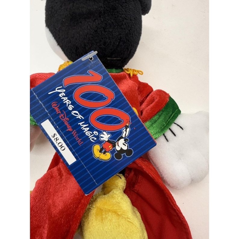 Puede incluir: Un peluche de Mickey Mouse con un atuendo rojo y verde. El juguete tiene una cabeza negra, guantes blancos y un torso amarillo. Una etiqueta azul dice "100 Years of Magic" y presenta a Mickey Mouse.