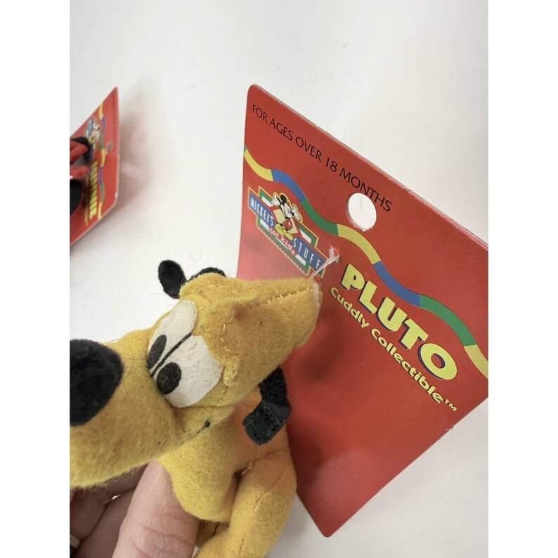 Puede incluir: Un peluche amarillo de Pluto con orejas y detalles negros, sujeto a una etiqueta de cart&oacute;n roja. La etiqueta dice "PLUTO Cuddly Collectible" y "For Ages Over 18 Months". El juguete es parte de la colecci&oacute;n Mickey's Stuff.