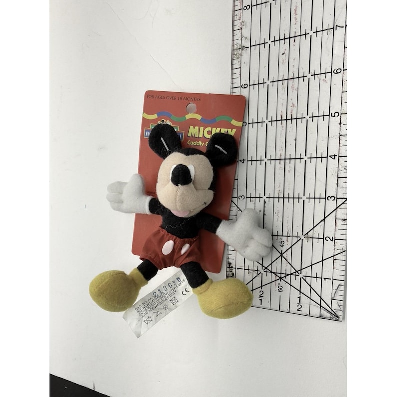 Puede incluir: Un peque&ntilde;o peluche de Mickey Mouse con orejas negras, cuerpo negro, guantes blancos, pantalones cortos rojos y zapatos amarillos. El juguete est&aacute; sujeto a una etiqueta de cart&oacute;n roja con la inscripci&oacute;n "MICKEY Cuddly".