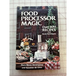 May include: A cookbook titled "FOOD PROCESSOR MAGIC" with the subtitle "Over 600 RECIPES and Basic Techniques." The cover features a food processor, various food items, and the authors' names: Mary Moon Hemingway and Suzanne de Lima.