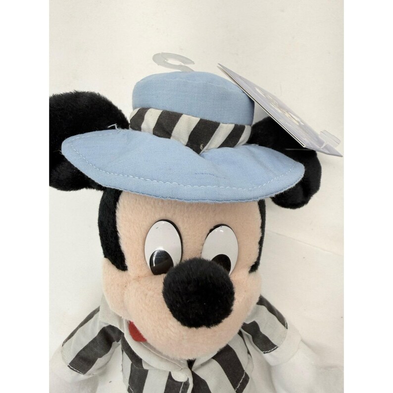 May include: A plush Mickey Mouse toy wearing a light blue hat with a black and white striped band. Mickey Mouse is wearing a black and white striped shirt. The toy has large white eyes and a black nose.