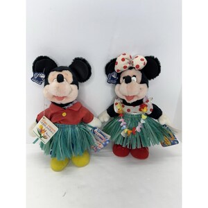 Puede incluir: Dos peluches de Disney, Mickey y Minnie Mouse, vestidos con atuendos hawaianos. Mickey lleva una camisa roja y una falda de hierba, Minnie un vestido de lunares y un lazo. Ambos tienen etiquetas.
