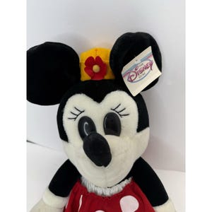 May include: A Minnie Mouse soft toy with black ears, a yellow and red flower crown, and a red dress with white polka dots. The toy has a tag from The Disney Store.
