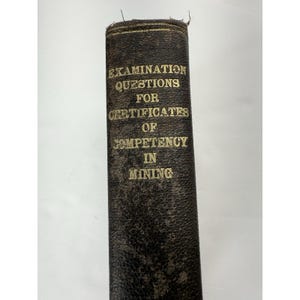 May include: A vintage book with a dark brown cover. The title "EXAMINATION QUESTIONS FOR CERTIFICATES OF COMPETENCY IN MINING" is printed in gold lettering. The book's spine shows signs of wear.