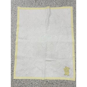 May include: A white baby blanket with a yellow gingham border and a small embroidered teddy bear in the corner. The blanket has a subtle dotted pattern across its surface, creating a soft and inviting texture.
