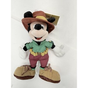 K&ouml;nnte beinhalten: Ein Mickey Mouse Pl&uuml;schtier mit einem braunen Hut mit gelbem Band, einem gr&uuml;nen Hemd, weinroten Hosen und braunen Schuhen. Das Pl&uuml;schtier hat wei&szlig;e Handschuhe und ein l&auml;chelndes Gesicht. Ein Disney-Etikett ist angebracht.