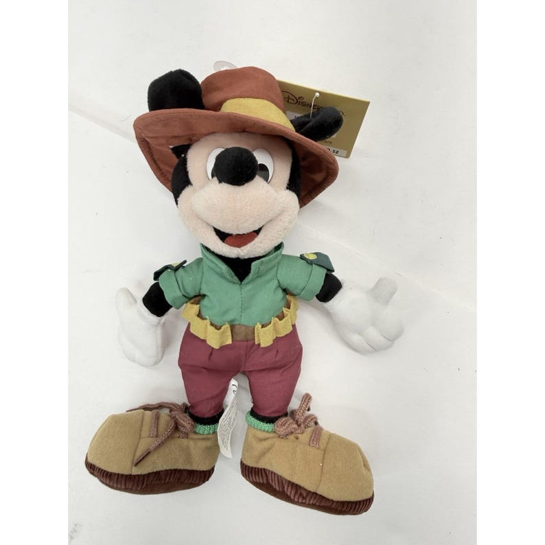 K&ouml;nnte beinhalten: Ein Mickey Mouse Pl&uuml;schtier mit einem braunen Hut mit gelbem Band, einem gr&uuml;nen Hemd, weinroten Hosen und braunen Schuhen. Das Pl&uuml;schtier hat wei&szlig;e Handschuhe und ein l&auml;chelndes Gesicht. Ein Disney-Etikett ist angebracht.