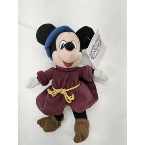 May include: Plush Mickey Mouse dressed in a burgundy robe with a gold rope belt and a blue hat. The stuffed animal has black ears, a pink face, and brown shoes. A Disney Store tag is attached.