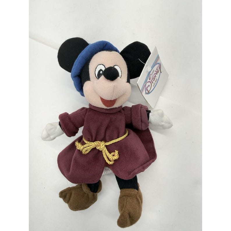 May include: Plush Mickey Mouse dressed in a burgundy robe with a gold rope belt and a blue hat. The stuffed animal has black ears, a pink face, and brown shoes. A Disney Store tag is attached.
