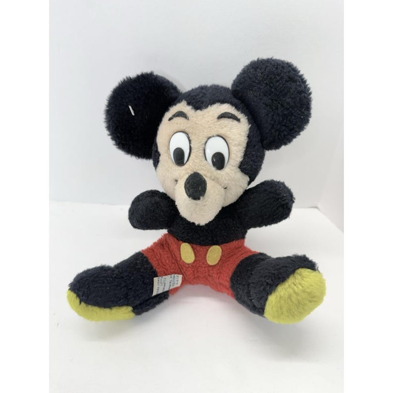 May include: A plush Mickey Mouse toy with black fur, large round ears, and a cream-colored face. He wears red shorts with yellow buttons and yellow-soled shoes. The toy is sitting with its legs spread.