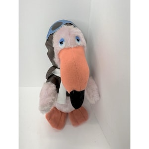 May include: A plush flamingo toy wearing a brown aviator jacket, blue goggles, and a white scarf. The flamingo has a large orange beak, pink body, and orange feet. The toy is designed to look like a pilot.
