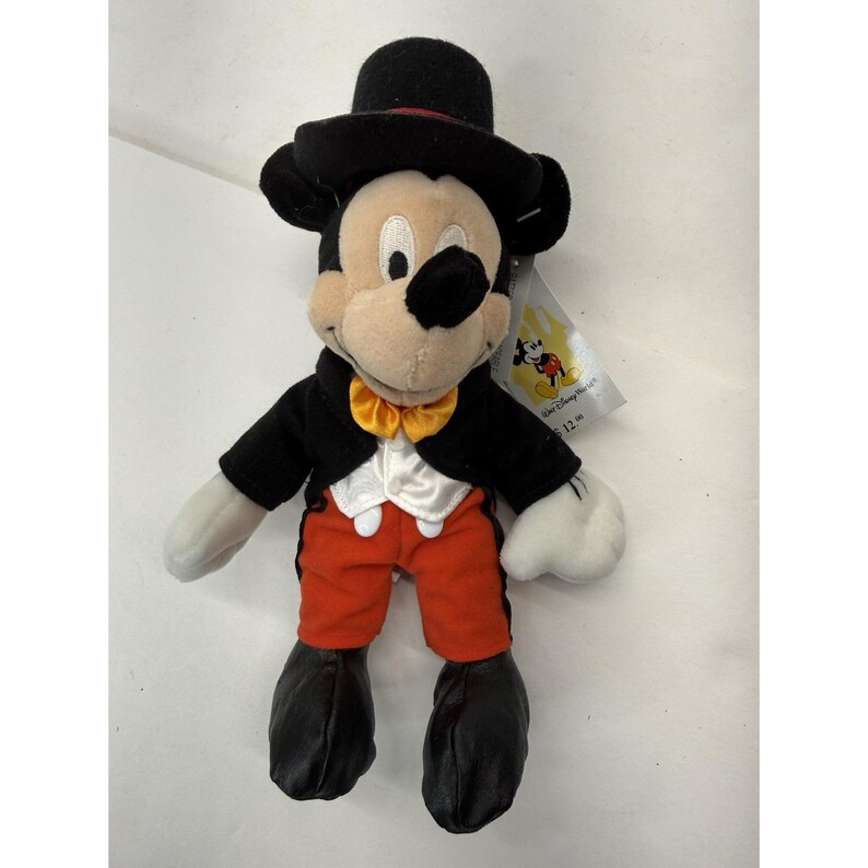 Peut inclure: Peluche de Mickey Mouse portant un chapeau haut de forme noir, une veste noire, un pantalon orange et un n&oelig;ud papillon jaune. Le jouet a une chemise et des gants blancs, avec des chaussures noires. Une &eacute;tiquette Disney est attach&eacute;e.