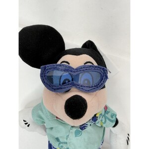 May include: A Mickey Mouse plush toy wearing blue sunglasses and a light green shirt with a floral pattern. The plush has black ears, a black nose, and white gloves. The toy is a Disney Store product.