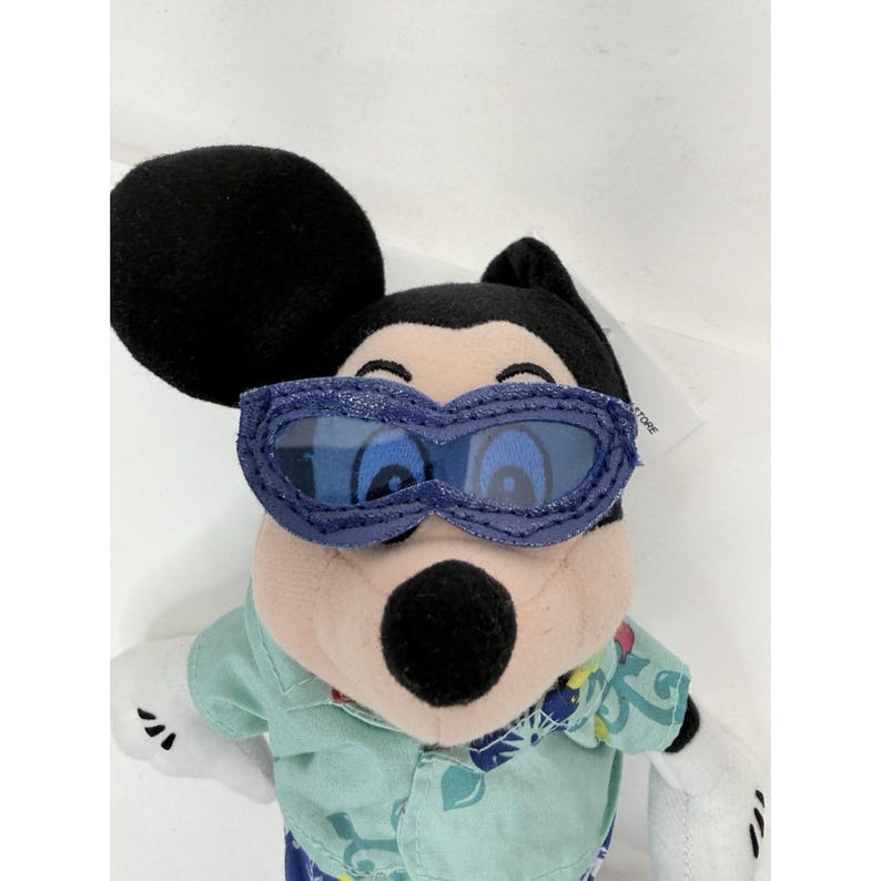 May include: A Mickey Mouse plush toy wearing blue sunglasses and a light green shirt with a floral pattern. The plush has black ears, a black nose, and white gloves. The toy is a Disney Store product.