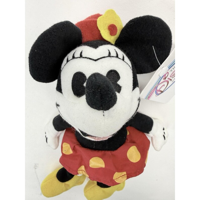 Puede incluir: Un mu&ntilde;eco de peluche de Minnie Mouse con orejas negras, una falda roja con lunares amarillos y zapatos amarillos. El mu&ntilde;eco tiene un sombrero rojo con una flor amarilla y una cara blanca. Una etiqueta de Disney est&aacute; adjunta.