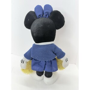 May include: A plush toy of a mouse character, viewed from the back. The mouse has black fur, large round ears, and a blue bow. It wears a blue sweater and shorts, white gloves, and white shoes. Yellow fur peeks out from the back.