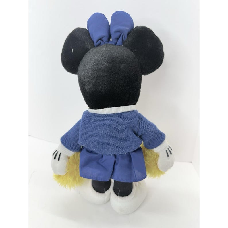 May include: A plush toy of a mouse character, viewed from the back. The mouse has black fur, large round ears, and a blue bow. It wears a blue sweater and shorts, white gloves, and white shoes. Yellow fur peeks out from the back.