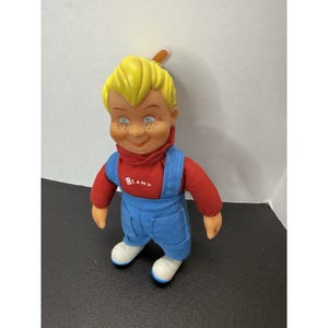 May include: A Beany doll with yellow molded hair, green eyes, and freckles on the face. The doll wears a red long-sleeved shirt with the word BEANY printed in white on the chest, blue denim-style overalls, and white shoes with blue trim.