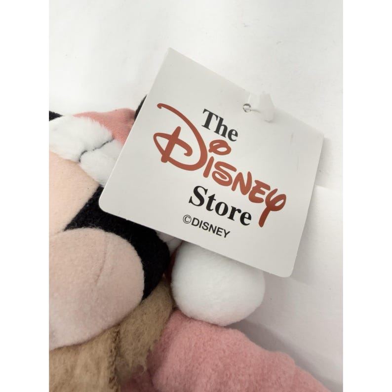 May include: A plush Minnie Mouse toy with a pink bow and dress, black ears, and a white pom-pom tail. The Disney Store tag is attached, featuring the Disney logo in red.