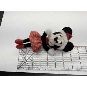 May include: A plush Minnie Mouse doll wearing a red polka dot skirt, red shoes, and a red and white hat. The doll has black ears, a black face, and white gloves. The doll is lying on its side.