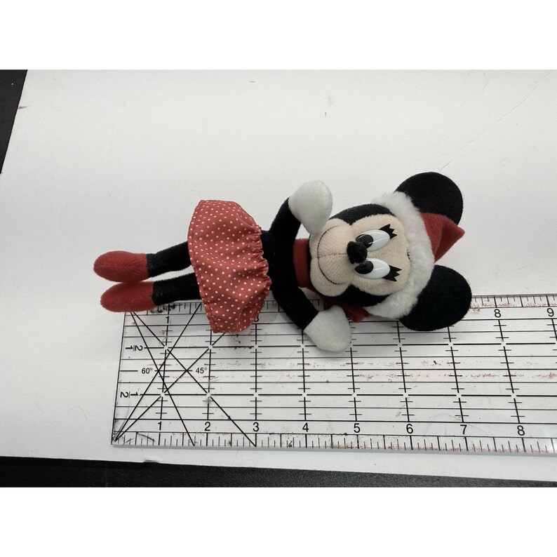 May include: A plush Minnie Mouse doll wearing a red polka dot skirt, red shoes, and a red and white hat. The doll has black ears, a black face, and white gloves. The doll is lying on its side.