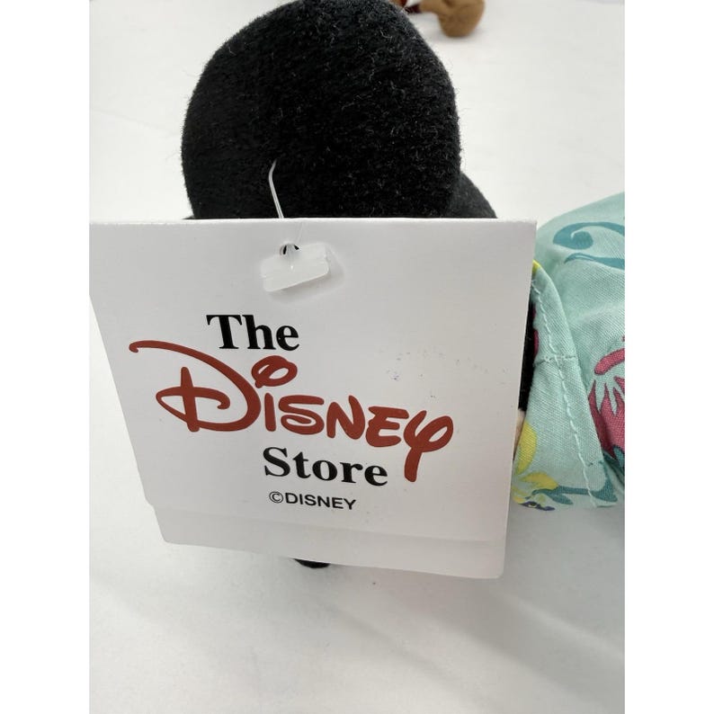 May include: A plush toy with a black head and a tag from The Disney Store. The tag is white with the Disney logo in red and the words "The Disney Store" in black. The toy has a light blue and floral patterned fabric.