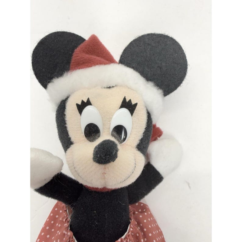 May include: A plush Minnie Mouse doll wearing a red and white Santa hat and a red polka dot dress. The doll has large, white eyes, black ears, and a black nose. The doll is a festive holiday decoration.