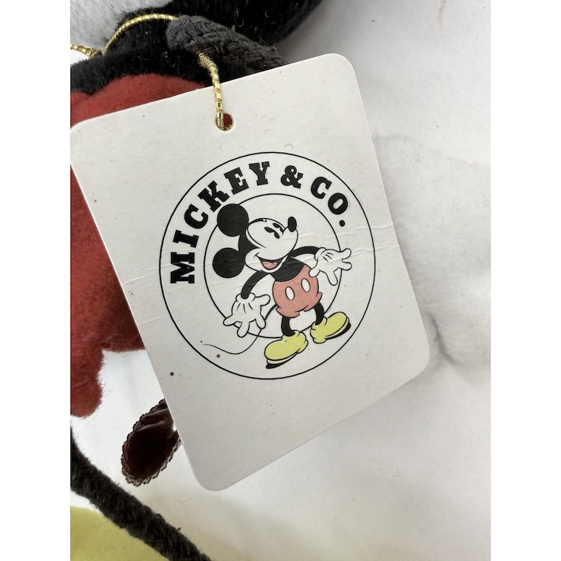 K&ouml;nnte beinhalten: Ein wei&szlig;es Etikett mit dem Text "MICKEY & CO." in einem kreisf&ouml;rmigen Design, das ein Cartoon-Bild von Mickey Mouse zeigt. Das Etikett ist an einem Pl&uuml;schtier mit roten, schwarzen und gelben Akzenten und einer goldenen Schnur befestigt.