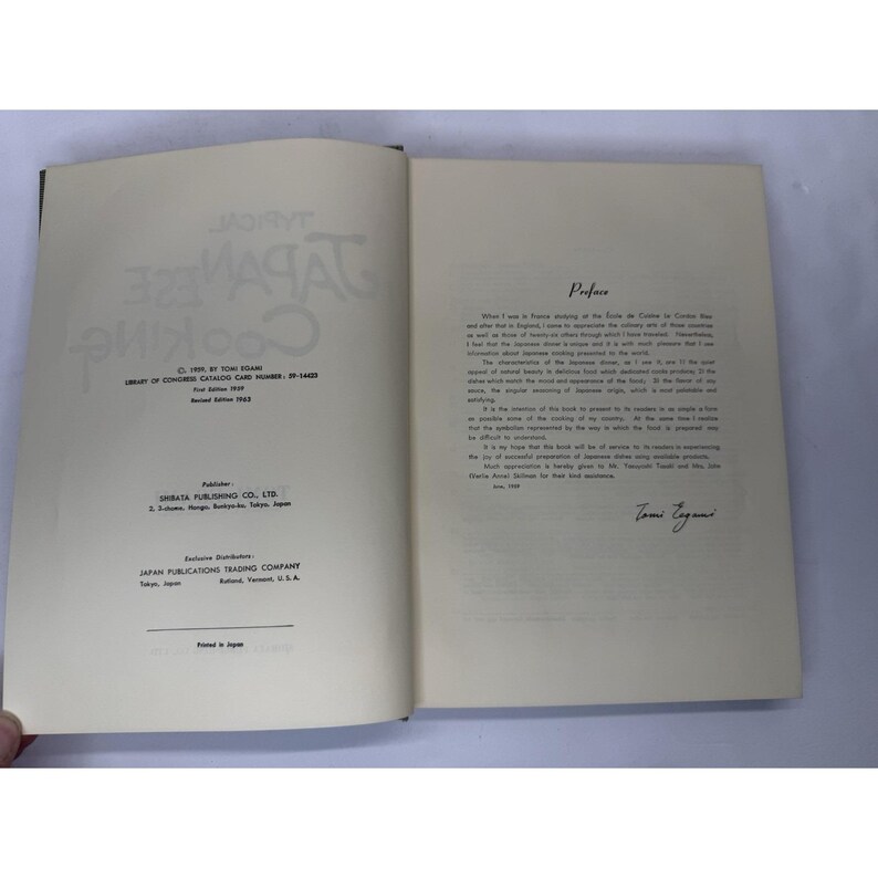 May include: An open book with the title "Typical Japanese Cooking" in large, stylized letters. The left page displays publishing information, while the right page features a preface. The book is cream-colored with black text.