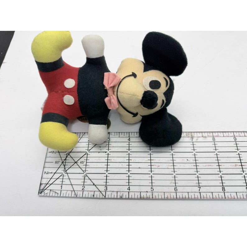 Puede incluir: Un peluche vintage de Mickey Mouse. El juguete est&aacute; en posici&oacute;n acostada, con una cabeza y orejas negras, una cara color crema y un lazo rosa. El cuerpo es rojo con botones blancos, pantalones cortos negros y pies amarillos. Una regla est&aacute; al lado del juguete.