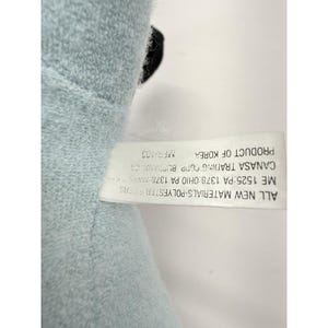 Puede incluir: Un peluche azul claro con un acento negro. Una etiqueta blanca adjunta muestra texto que incluye "ALL NEW MATERIALS-POLYESTER FIBERS" y "PRODUCT OF KOREA".