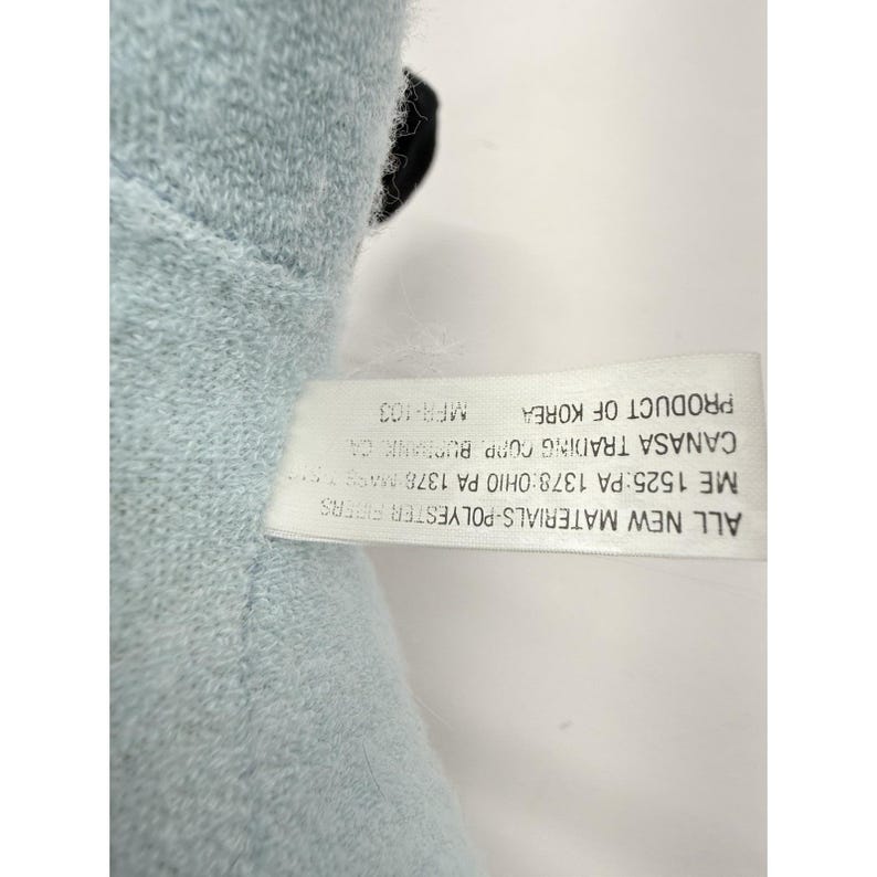 Puede incluir: Un peluche azul claro con un acento negro. Una etiqueta blanca adjunta muestra texto que incluye "ALL NEW MATERIALS-POLYESTER FIBERS" y "PRODUCT OF KOREA".