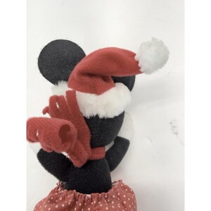 May include: A plush Minnie Mouse figure wearing a red Santa hat with a white pom-pom, a red scarf, and a red polka-dot skirt. The figure has black ears and a black body, perfect for holiday decor.
